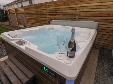 An outdoor hot tub with bubbling water two empty wine glasses and a bottle of prosecco on the edge near a wooden fence at Sea Cliff in Morfa Nefyn