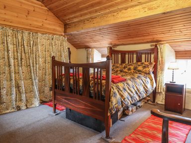 A bedroom with a wooden bed, floral curtains, a nightstand with a lamp, and red rugs at The Long House in Penmachno