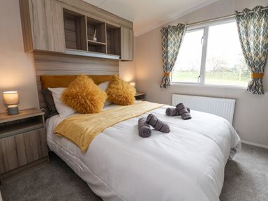 A bedroom with a double bed yellow pillows and blanket bedside tables with lamps and a window with patterned curtains at Lazy Dayz Lodge East Heslerton near Malton