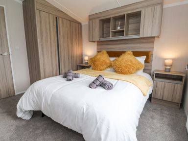 A bedroom with a double bed white bedding yellow pillows rolled towels wooden wardrobes and bedside tables with lamps at Lazy Dayz Lodge in East Heslerton near Malton