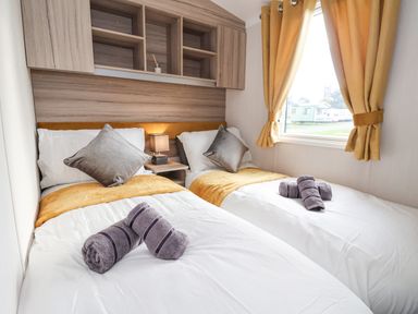 A bedroom with two single beds with grey pillows and rolled towels by a window with yellow curtains at Lazy Dayz Lodge East Heslerton near Malton