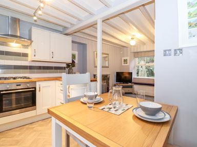 A kitchen with a wooden table set with dishes and a glass jug next to a living area with a television and window seat at Fell View in Ambleside