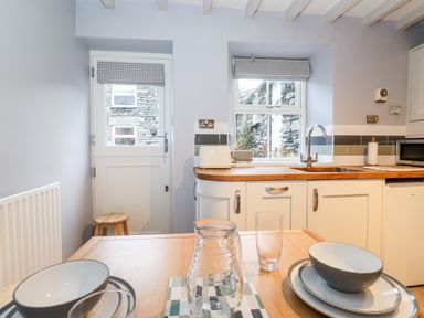A kitchen with a wooden table set with bowls plates glasses and a jug with a sink microwave toaster and window in the background at Fell View in Ambleside
