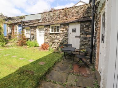 A stone cottage with white doors and windows a small patio with a table and two chairs and a grassy garden at Fell View in Ambleside