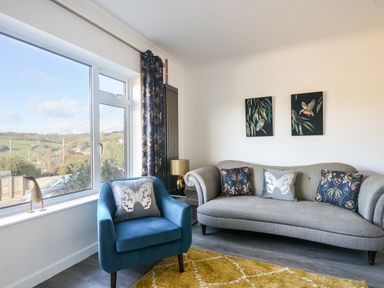 A living room with a gray sofa and a blue armchair near a large window with patterned curtains at Hook Farm Cottage in Uplyme