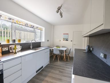 A kitchen with white cabinets gray countertops a sink window with patterned blind and a dining table with chairs at Hook Farm Cottage in Uplyme