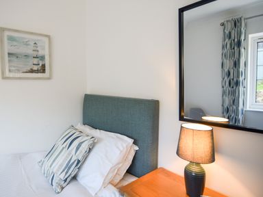 A bedroom corner with a bed, patterned pillow, a wooden nightstand with a lamp, a mirror, and a framed picture of a lighthouse at Hook Farm Cottage in Uplyme