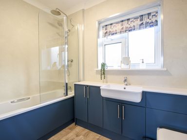 A bathroom with a bathtub shower combination and sink with blue cabinets under a window at Hook Farm Cottage in Uplyme