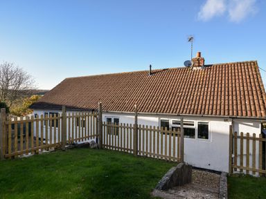 A white house with a tiled roof behind a wooden fence and grass at Hook Farm Cottage in Uplyme