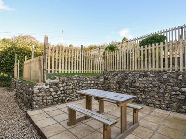 An outdoor patio area with a wooden picnic table and benches surrounded by stone walls and wooden fencing at Hook Farm Cottage in Uplyme