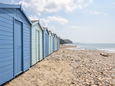 A row of blue beach huts on a sandy and pebbly shore by the sea with cliffs in the distance at Hook Farm Cottage in Uplyme