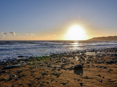 A sandy and rocky beach with ocean waves and the sun setting near a coastal hill