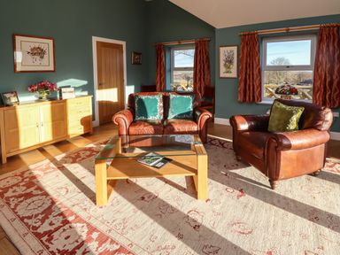 A living room with leather armchairs wooden coffee table sideboard and windows with red curtains at Summerseats Cottage in Alnwick