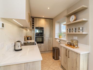 A kitchen with wooden cabinets a kettle a toaster built in ovens shelves with a clock and containers at Summerseats Cottage in Alnwick