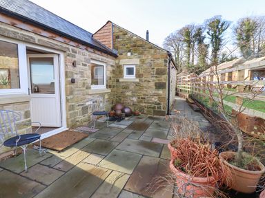 An outdoor stone patio with metal chairs and potted plants adjacent to a stone cottage at Summerseats Cottage in Alnwick