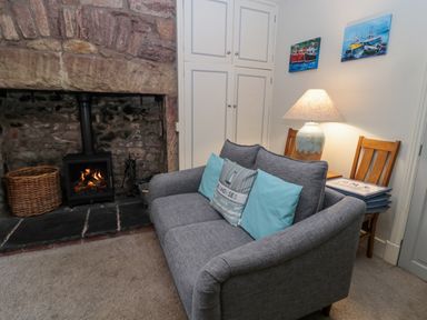 A living room with a gray sofa near a stone fireplace with a fire and a lamp on a table at The Haven in North Sunderland near Seahouses
