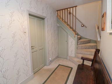A hallway with stairs a closed door a small door under the stairs a bench and wallpaper with branch patterns at The Haven in North Sunderland near Seahouses
