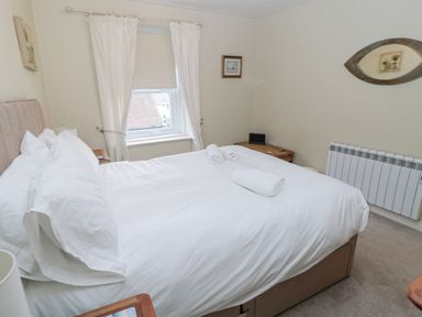 A bedroom with a large bed made with white sheets and rolled towels on top two wooden side tables a window with white curtains and a wall mirror at The Haven in North Sunderland near Seahouses