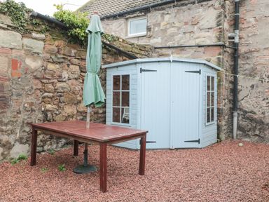 An outdoor area with a red gravel ground a brown wooden table with a closed green umbrella stone walls and a light blue shed at The Haven in North Sunderland near Seahouses