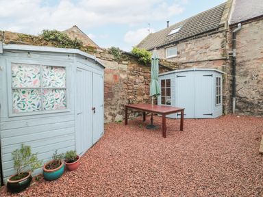 An outdoor area with two blue sheds a brown table with a closed umbrella and three potted plants on gravel at The Haven in North Sunderland near Seahouses
