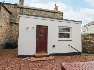 A small white building with a brown door and a window next to a stone wall at The Haven in North Sunderland near Seahouses