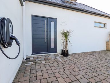 A paved outdoor area with a dark blue door a potted plant and an electric vehicle charger on a white wall at Little Beach Cabin in Crantock