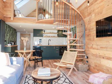 A living room with wooden walls spiral staircase and kitchen area at Little Beach Cabin in Crantock