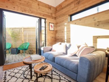 A living area with a gray sofa and pillows a patterned rug and nested wooden tables opening to a patio with green chairs at Little Beach Cabin in Crantock