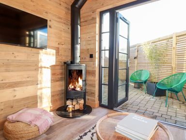 A room with a wood stove burning next to an open door leading to a patio with two green chairs at Little Beach Cabin in Crantock