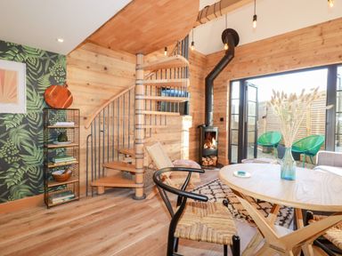 A living room with a spiral staircase a wood stove a round wooden table and green chairs outside at Little Beach Cabin Crantock