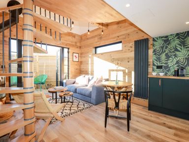 An open living space with a spiral staircase wooden walls a blue sofa round wooden tables a rug and a kitchenette with green cabinets and plant patterned wallpaper at Little Beach Cabin in Crantock