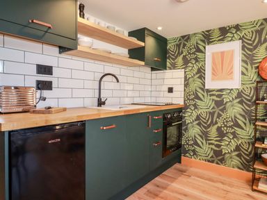 A kitchen with dark green cabinets wooden countertops white subway tile backsplash and patterned wallpaper at Little Beach Cabin in Crantock