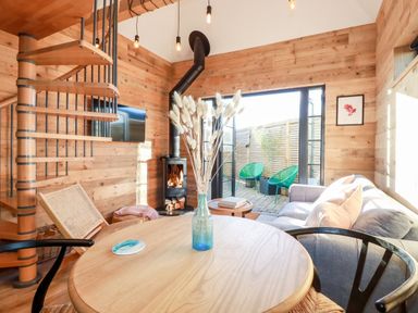 A wooden living room with a round table holding a vase with dried flowers a spiral staircase a wood stove and a gray sofa at Little Beach Cabin in Crantock