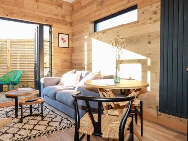 A living room with a gray sofa wooden tables a round wooden dining table and chairs with an open door leading outside at Little Beach Cabin in Crantock