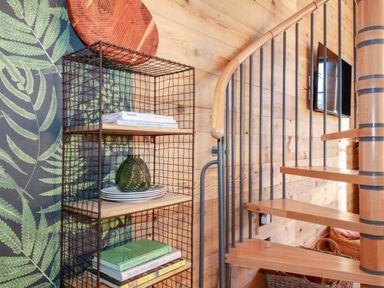 A wire shelving unit with books plates and a decorative item next to a wooden spiral staircase in a room with wood panel walls and fern wallpaper at Little Beach Cabin in Crantock