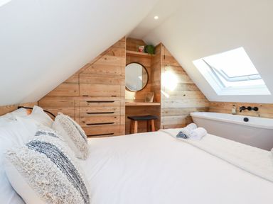 A bedroom with a bed, pillows, wooden panel walls, a bathtub under a skylight, and a small vanity with a round mirror at Little Beach Cabin in Crantock