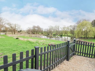 A black wooden fence with a gate overlooking a grassy area and a pond with trees at The Hayloft in Haddiscoe