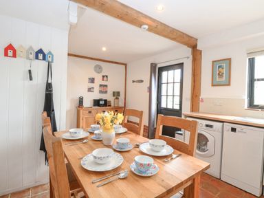 A dining area with a wooden table set with dishes and cups a sideboard with a microwave and lamp a door leading outside and kitchen appliances at The Hayloft Haddiscoe