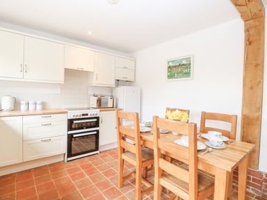 A kitchen with wooden dining table and chairs set for tea beside white cabinets with stove and refrigerator at The Hayloft in Haddiscoe