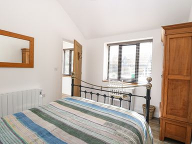 A bedroom with a striped bedspread a wooden wardrobe a white chair and desk a window with blinds and a wooden mirror at The Grain Store in Haddiscoe