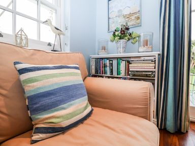 A living room corner with a beige sofa and striped cushion near a bookcase with books flowers and candles at Lampland House in Walberswick
