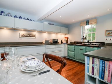 A kitchen with white cabinets a green stove a table set with plates and glasses and a bookshelf with cookbooks at Lampland House in Walberswick