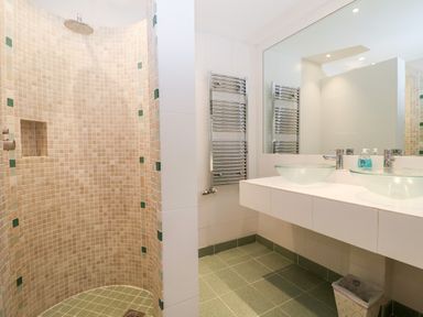 A bathroom with a curved tiled shower, double glass sinks, a large mirror, a towel warmer, and a trash bin at Lampland House in Walberswick