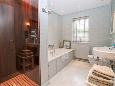 A bathroom with a bathtub a shower with glass door a sink a toilet folded towels on a stool and a window with blinds at Lampland House in Walberswick