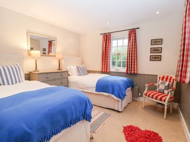 A bedroom with two single beds blue blankets striped pillows a dresser with two lamps a window with red check curtains and a striped armchair at Lampland House in Walberswick