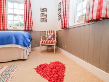 A bedroom with a bed covered by a blue blanket a chair with red and white stripes red checkered curtains and a red heart shaped rug on the floor at Lampland House in Walberswick