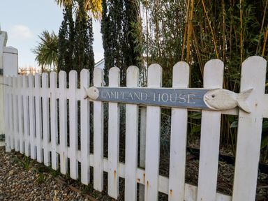 A white wooden picket fence with a sign reading Lampland House attached to it with trees and plants behind it at Lampland House in Walberswick