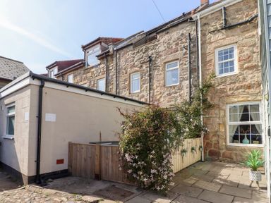 An outdoor courtyard area with stone paving surrounded by stone and plaster building walls with windows and climbing plants at Seascape Cottage in Seahouses