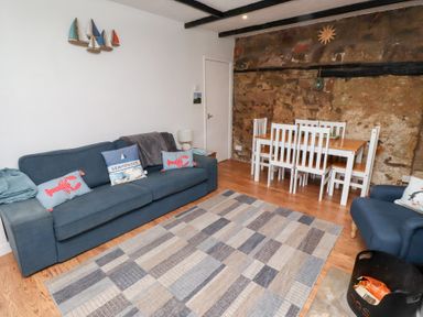 A living room with a blue sofa a dining table with white chairs and a patterned rug at Seascape Cottage in Seahouses
