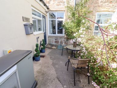 A small outdoor patio area with two chairs and a round glass table next to a wall covered with flowering plants at Seascape Cottage in Seahouses
